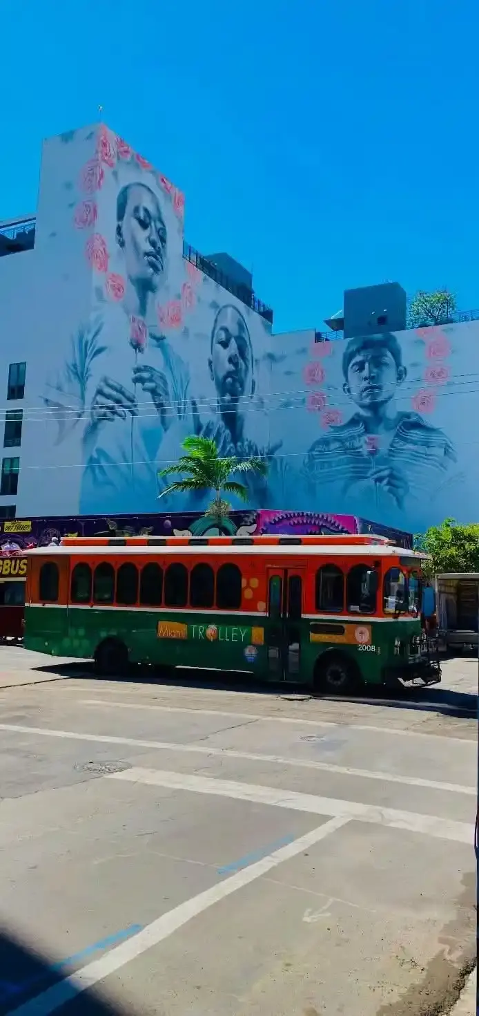 Un trolley orange et vert de la ville de Miami stationné dans une rue devant un grand bâtiment orné d'une fresque murale artistique en noir et blanc représentant trois visages entourés de roses.