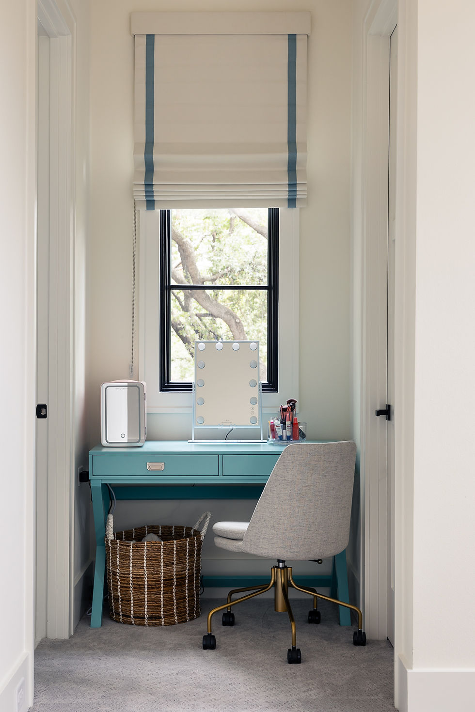 Makeup vanity with lighted mirror on a teal desk, beige chair, and wicker basket. Window with blue-striped shade. Bright, cozy setting.