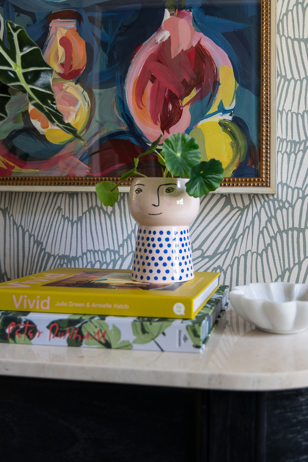 Ceramic vase with blue polka dots holding leaves, placed on colorful books. Abstract painting and patterned wallpaper in the background.
