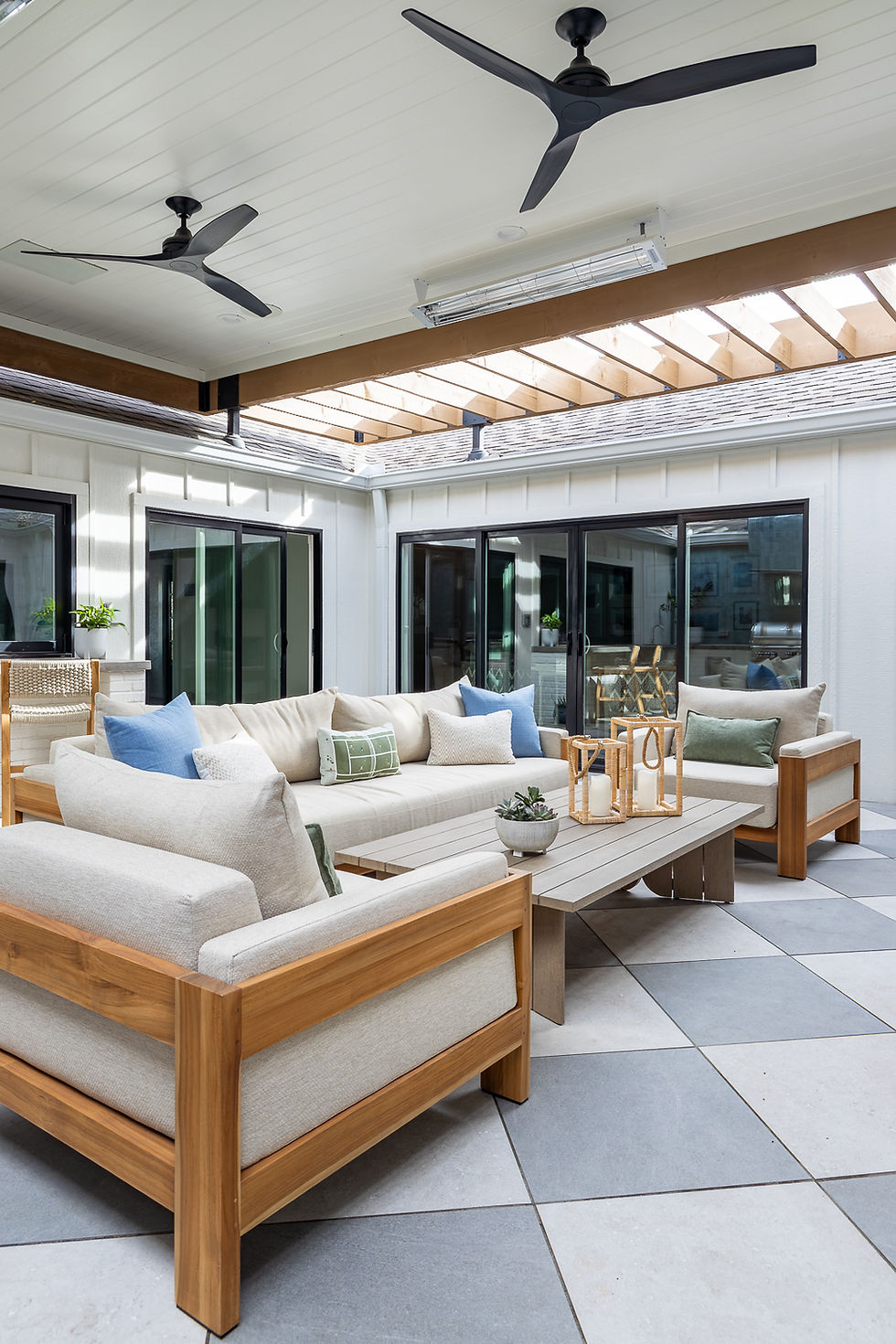 Outdoor patio with beige wooden furniture, blue and green cushions, and candle lanterns on a table. Ceiling fans above and natural light.