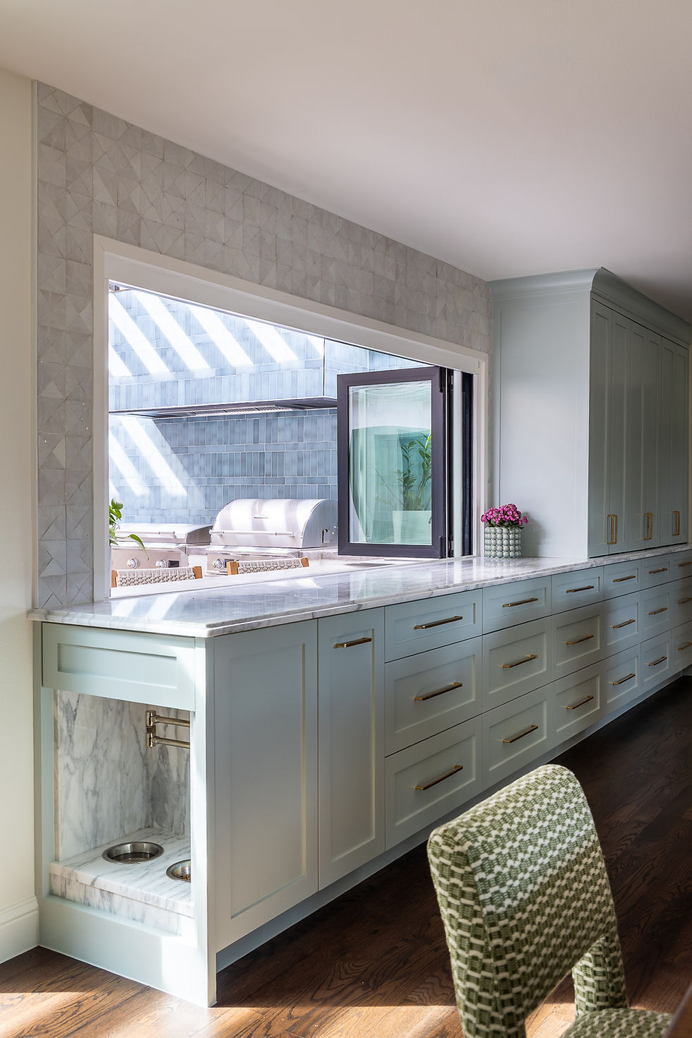 Spacious kitchen with light blue cabinets and marble countertop. Open window reveals outdoor grill. Sunlight streams in, creating a calm mood.