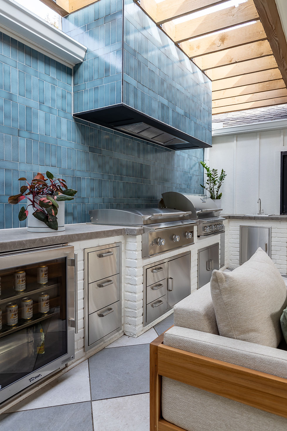 Outdoor kitchen with stainless steel grill, fridge, and blue tiled wall. Wooden ceiling lets in light, and potted plants add greenery.