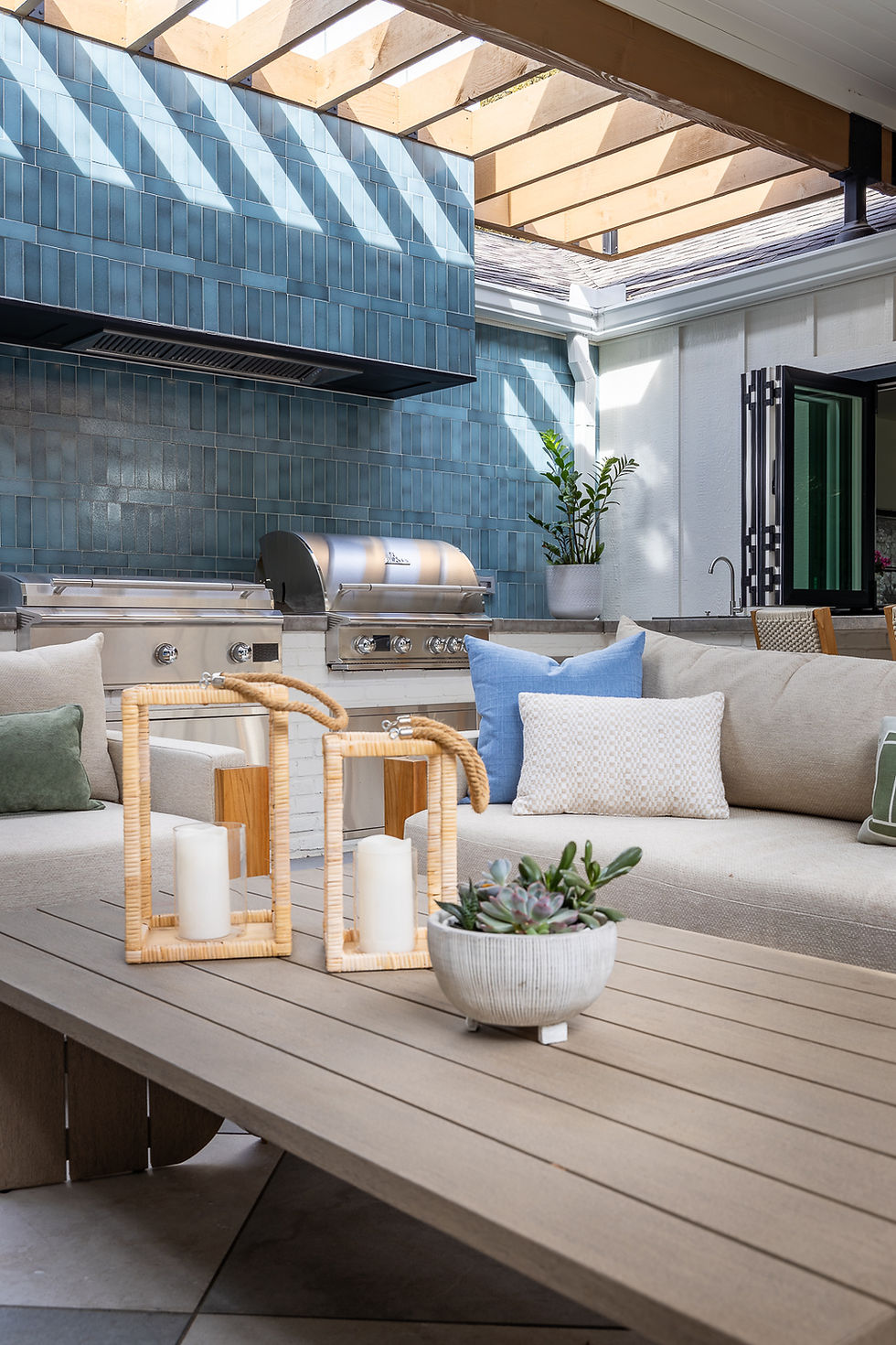 Outdoor seating area with beige sofa, colorful pillows, and candles on a wooden table. Blue tiled wall and grill in the background.