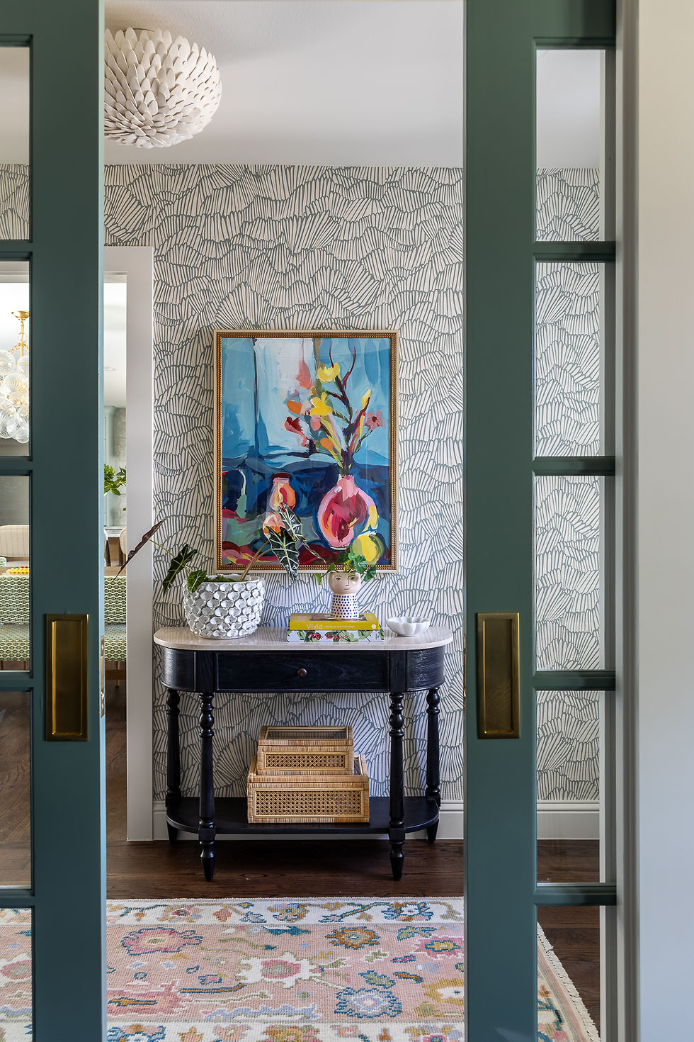 Artistic hallway with abstract painting above a black table, plants, books, and woven baskets. Colorful rug and patterned wallpaper.