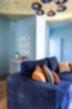 Blue room with a velvet sofa, colorful pillows, and geometric ceiling. Art on the wall above a console with books and a plant.