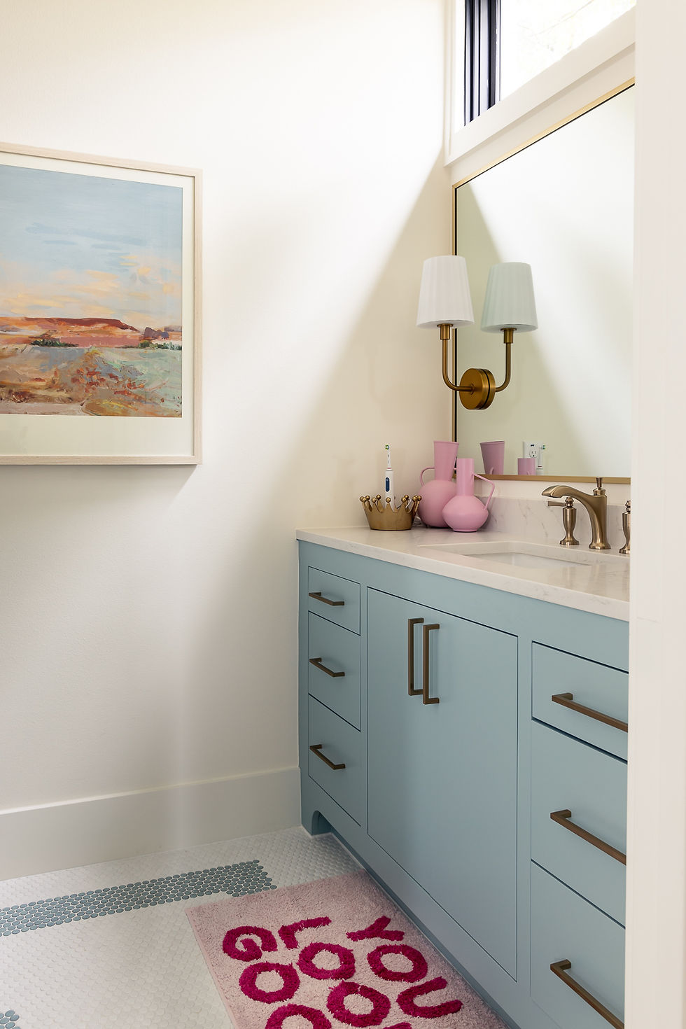 A bathroom with a light blue vanity, brass hardware, pink vases, and a framed landscape painting. Rug reads "you look good."