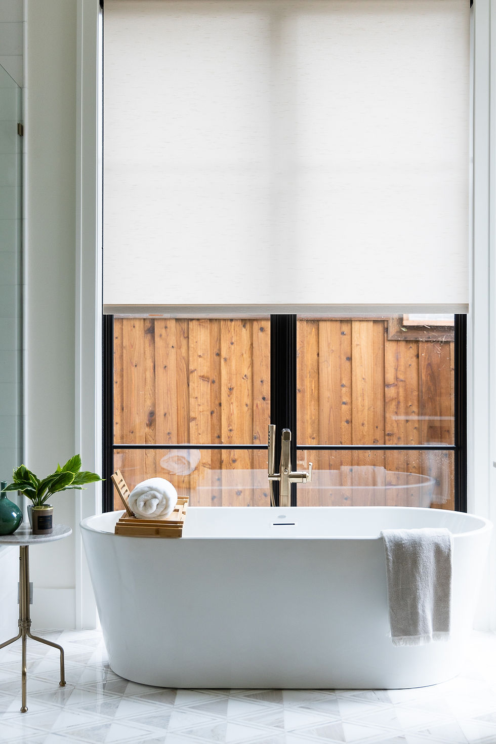 Modern bathroom with a white freestanding tub, rolled towel on a tray, and a wooden window view. Potted plant on a table; serene atmosphere.
