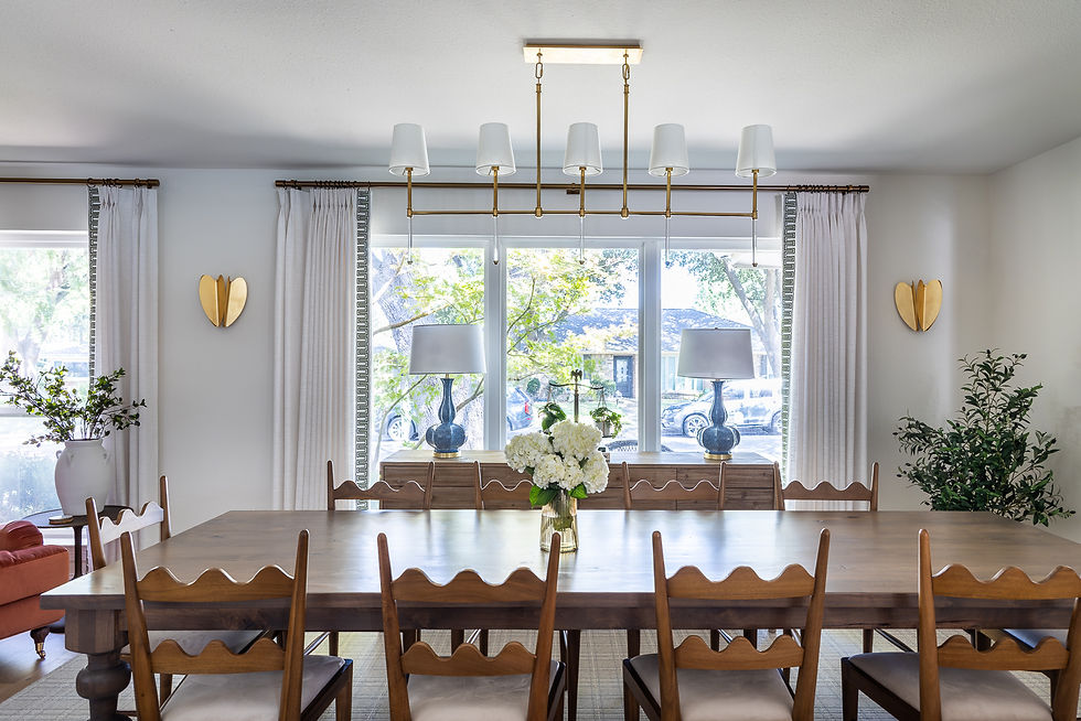Dining room with a long wooden table and chairs, white curtains, large window, two blue lamps, and a vase of white flowers under a chandelier.