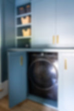 Blue laundry room with a black washing machine behind open cabinet doors. Shelves hold woven baskets. Gold handles accent the cabinets.