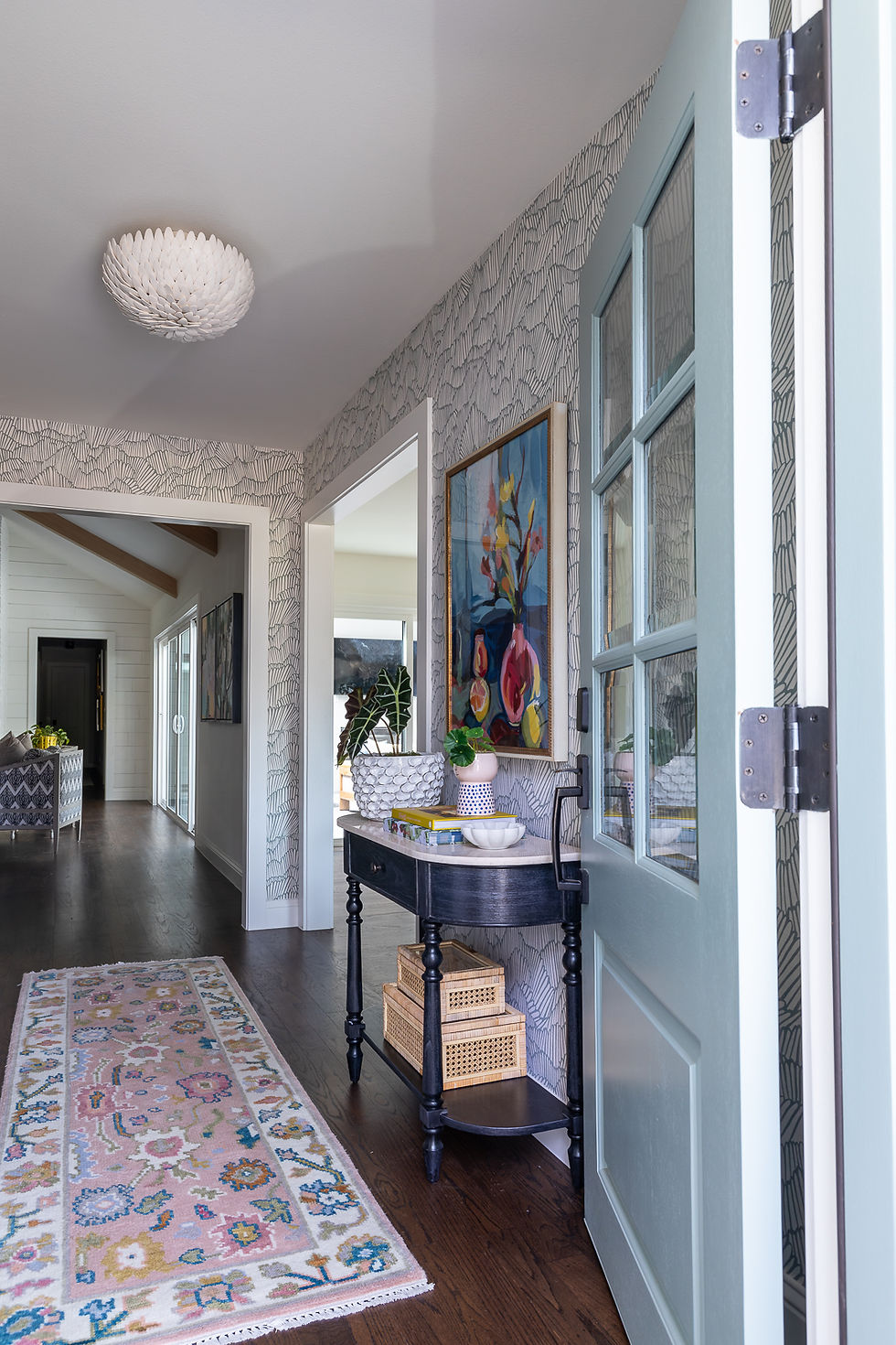 Hallway with patterned wallpaper, a light blue open door, floral painting, and a console table with decor. Colorful rug on wood floor.