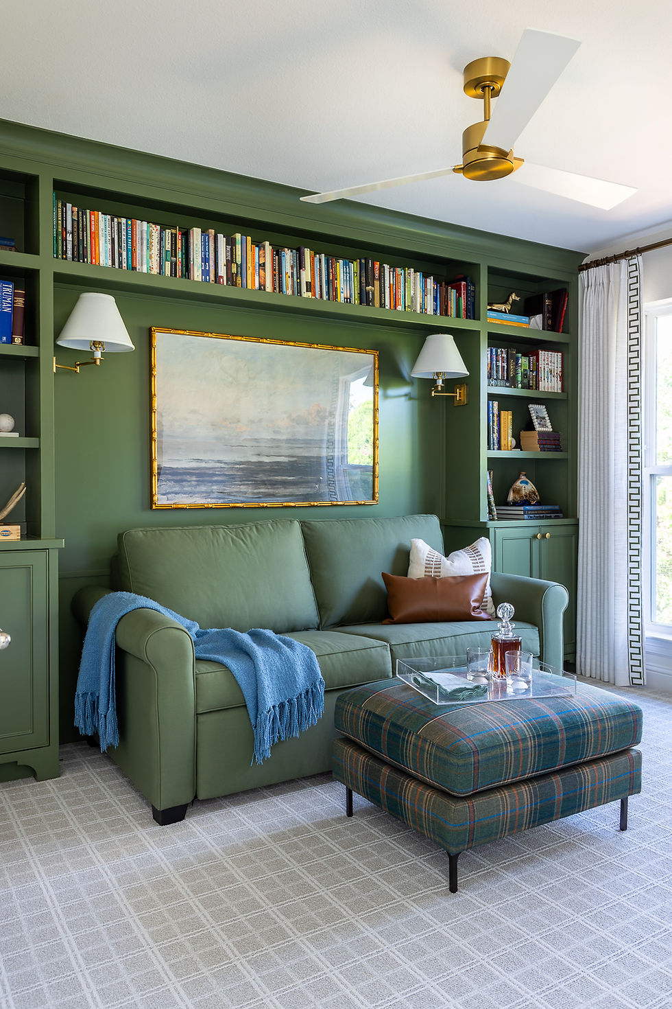 Cozy room with green sofa, blue throw, plaid ottoman, and bookshelves. Beach art, glass decanter, gold fan, and light create a serene vibe.