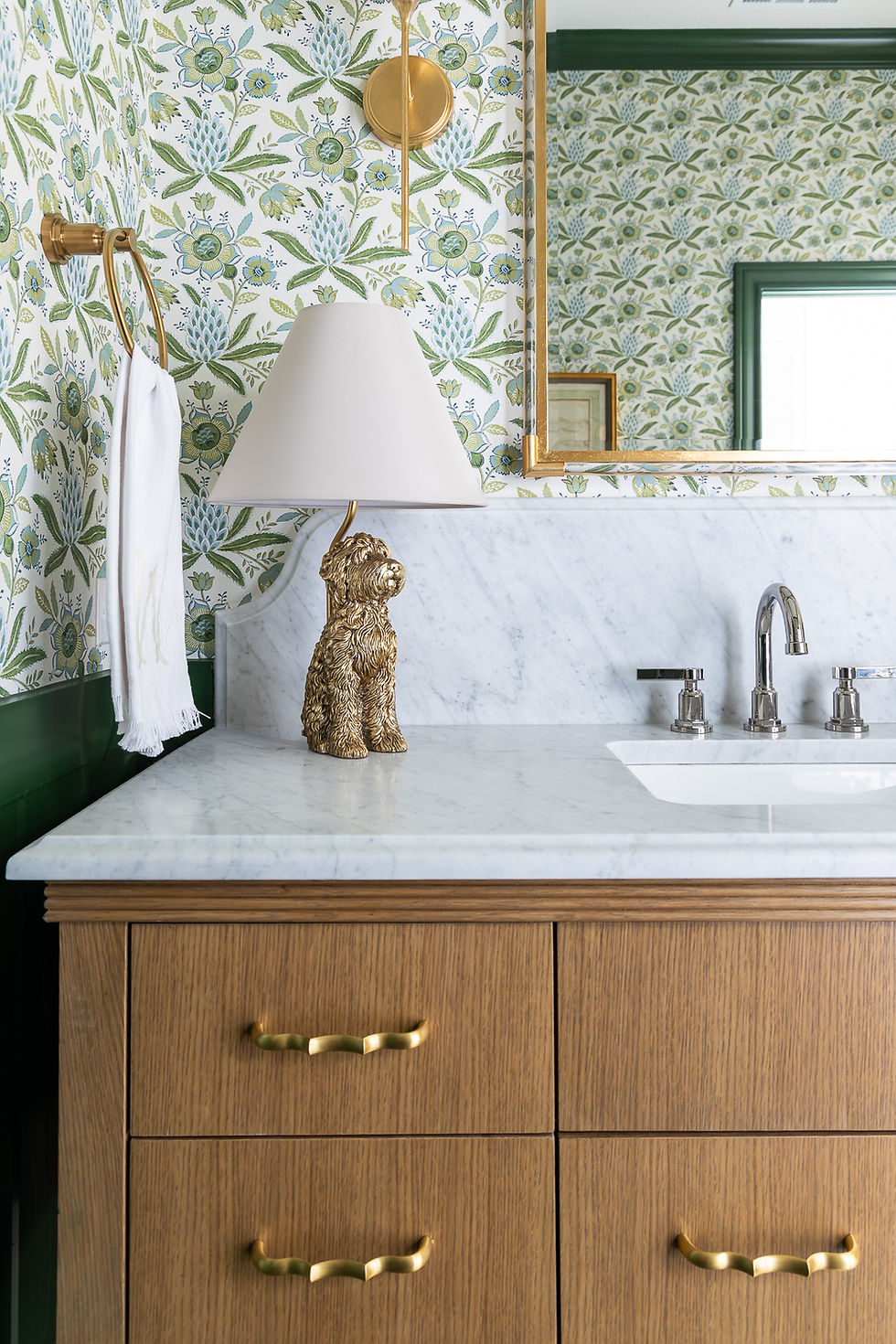 Bathroom vanity with marble top, golden dog lamp, floral wallpaper, gold fixtures, and wooden drawers. Calm and stylish setting.