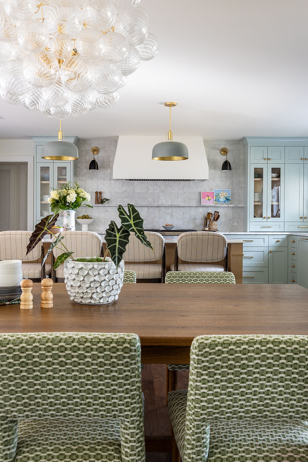 Elegant kitchen with green pattern chairs, wooden table, and a textured plant pot. Soft lighting, mint cabinets, and a spherical chandelier.