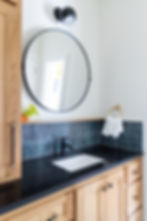 Modern bathroom vanity with a round mirror, black fixtures, dark countertop, wooden cabinetry, and a potted plant. Navy tile backsplash.