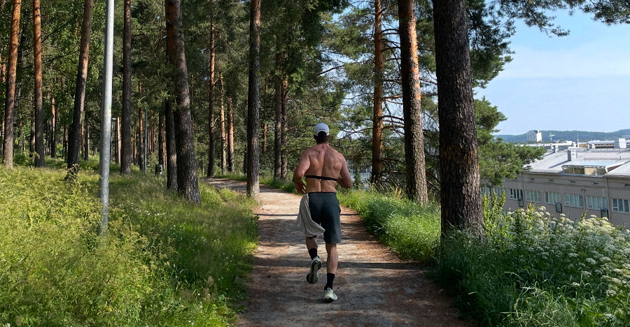 Man jogging on a wooded path, wearing shorts and a cap. Sunny day with buildings visible in the distance, conveying a peaceful mood. Hybrid athlete