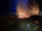 A nighttime fire burns a wooden fence and trees, casting an orange glow. Smoke billows, surrounded by a dark yard and scattered plants.