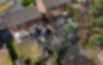 Aerial view of a burned-down backyard next to intact houses, with two people inspecting the charred remains amid green trees and lawns.