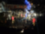 Firefighters in red jackets extinguish a fire at a charred house at night. Smoke and bright lights fill the scene, trees partly visible.