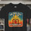 Thumbnail: BLACK TEE SHIRT WITH A COLORFUL PRINTED DESIGN WITH A GLOWING SUNSHINE BEHIND A CROSS AND A GIFT PACKAGE