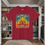 Thumbnail: RED TEE SHIRT WITH A COLORFUL PRINTED DESIGN WITH A GLOWING SUNSHINE BEHIND A CROSS AND A GIFT PACKAGE