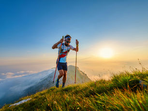 A man hiking uphill on a grassy mountain ridge at sunrise, using trekking poles for support, surrounded by clouds and golden sunlight — symbolizing perseverance, focus, and progress toward goals.