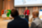 Woman (Dr Gali Artzi) in red jacket presents at a conference, holding a remote, with audience in foreground and slides on nutrition economy.