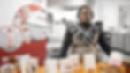 Woman stands by a table with Root Mylk brochures and products in the Medway Food Innovation Centre lab. Wears patterned dress and glasses, looking at the camera.