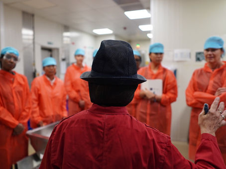 Person (Nimisha Raja MBE) in a hat lectures a group in orange coats and blue caps in a food factory. Focus on attentive expression and industrial setting.