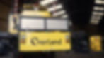 Yellow industrial machine with "Overland" text in a warehouse. Control buttons visible. Background shows striped poles and lit ceiling windows.