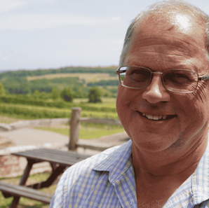 Robert Mitchell, wearing glasses and light check shirt, stands in front of rolling green hills