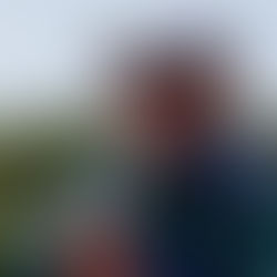 Man (Richard Small) in sunglasses and cap smiling outdoors, wearing a green jacket. Background of trees and cloudy sky suggests a cheerful nature setting.