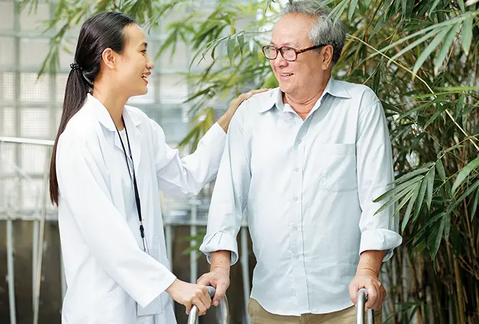 A healthcare professional supporting her elderly patient who is standing using a walker.