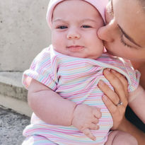 Mia kisses her baby daughter, who is wearing a pink hat and striped onesie.