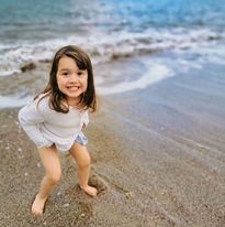 Mia's daughter smiles at the camera, squatting on the sandy beach with waves in the background.