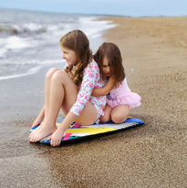 Two young girls sitting on a surfboard on the beach, the older one embracing the younger.