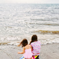 Mia's daughters sit on a bodyboard by the sea, looking out into the distance.