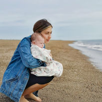 Mia crouches on the beach, holding her baby daughter, with the sea in the background.