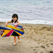 Mia's young daughter stands on the beach holding a colourful bodyboard, with wind turbines in the distance.