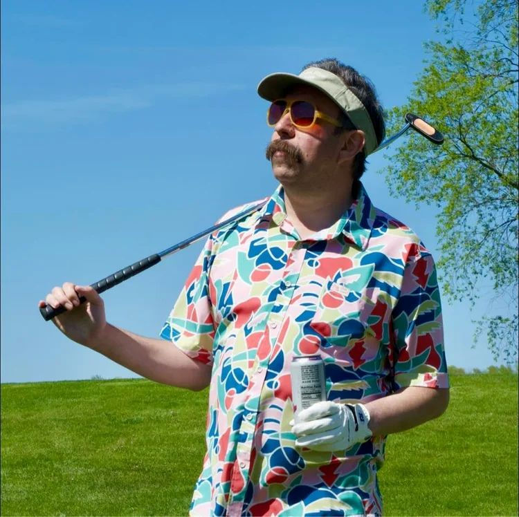 Joshua Sutton, owner of Trailyard Outpost, stands outdoors on a grassy field holding a golf club over his shoulder and a drink in his hand. He is wearing a colorful patterned shirt, sunglasses, a visor, and a golf glove, with a blue sky and tree in the background.