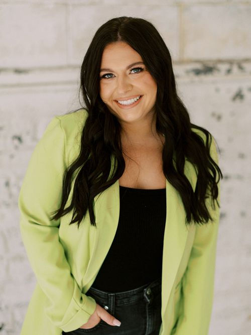 Logan Montague, owner of Socially Logan Marketing, smiling and standing against a light-colored textured wall. She is wearing a bright green blazer over a black top, with long dark hair and a confident, friendly expression.