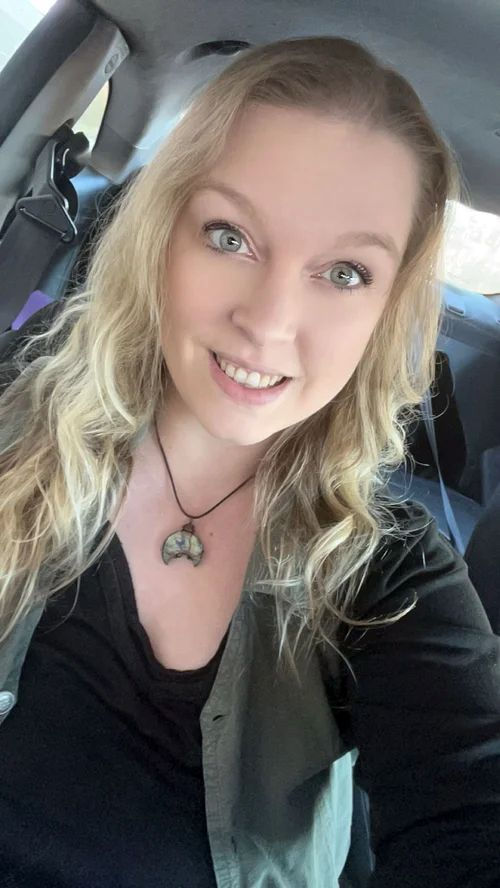 Brittany Goers, owner of Break the Mold Hypnosis, smiles at the camera while sitting in a car. She has long wavy blonde hair, wears a black top with a green shirt over it, and a crescent-shaped pendant necklace.