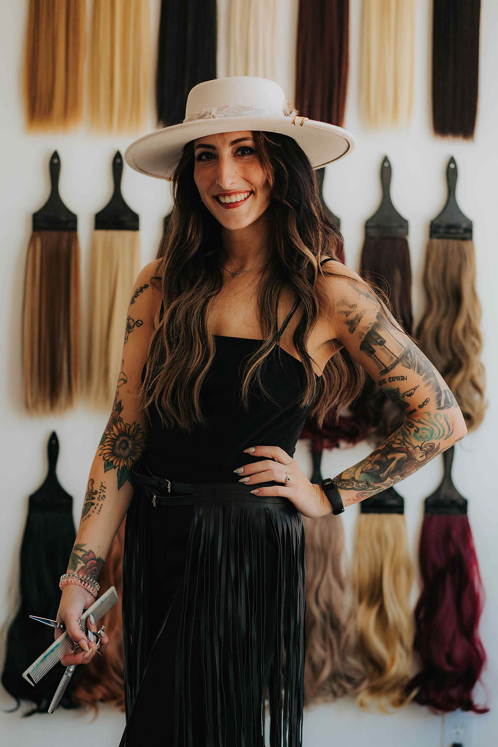Lauren Peters, owner of Fringe + Ink Creative Co, smiling and standing in front of a wall display of hair extensions. She has long wavy hair, wears a wide-brimmed hat and a black outfit with fringe, and has visible tattoos on her arms.