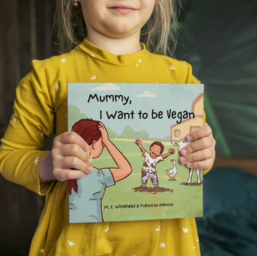 Child in a yellow dress holds a book titled Mummy, I Want to Be Vegan, with farm animals on the cover.