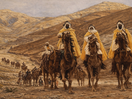 Desert caravan with camels and riders in yellow cloaks, moving through rocky terrain. Background features arid hills under a clear sky.