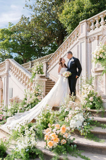 The couple pose on the stairway fully adorned with flowers, framed by soft blooms that enhance the elegance and romance of the setting