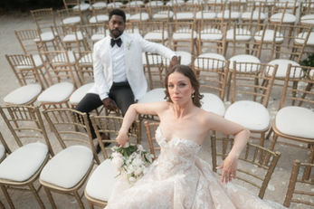 Bride seated among ceremony chairs holding a bouquet, with the groom behind her, during their wedding at Forte da Cruz in Cascais