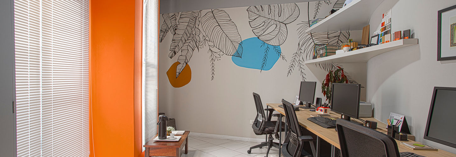 Sala de produção do escritório com bancada linear de madeira, cadeiras ergonômicas e parede decorada com mural artístico botânico e coluna laranja vibrante.