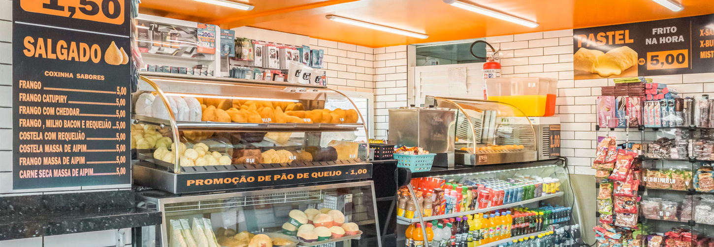 Detalhe do balcão refrigerado e expositor de salgados, pães e pão de queijo, com prateleiras de vidro e revestimento de fundo em azulejo branco.