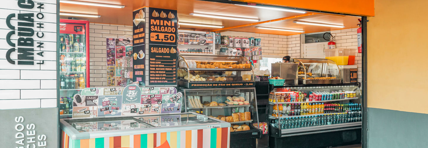 Vista de dentro do quiosque, mostrando o balcão principal com exposição de salgados, bebidas e logo vertical na coluna de azulejo branco.