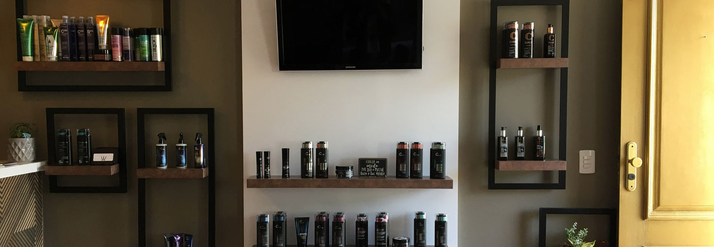 Parede de exposição de produtos do salão, com prateleiras flutuantes em metal preto e madeira, centralizadas por uma televisão.